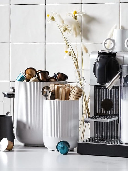 White cylindrical containers with coffee accessories next to a coffee machine on a tiled wall background