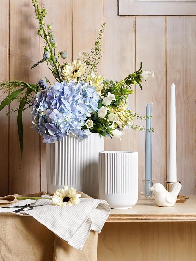 Floral arrangement in a white vase on a wooden surface with candles and a bird figurine.